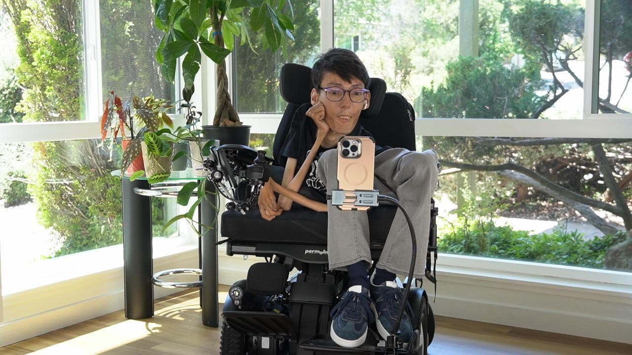 Vahid Rajabloo, an accessibility technology builder, seated in his power wheelchair with a phone mounted in front of him, in a bright, plant-filled room.